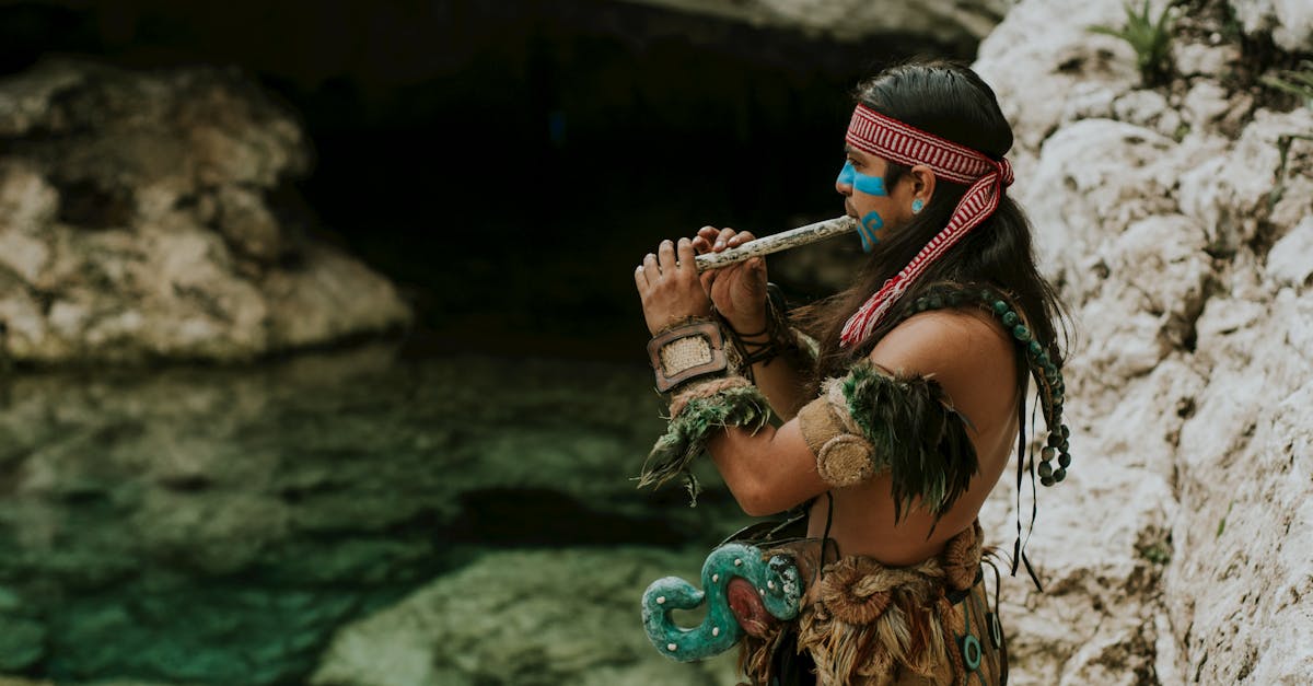 A Mayan musician wearing traditional attire plays a flute by a rocky water landscape, exuding cultural heritage and art.