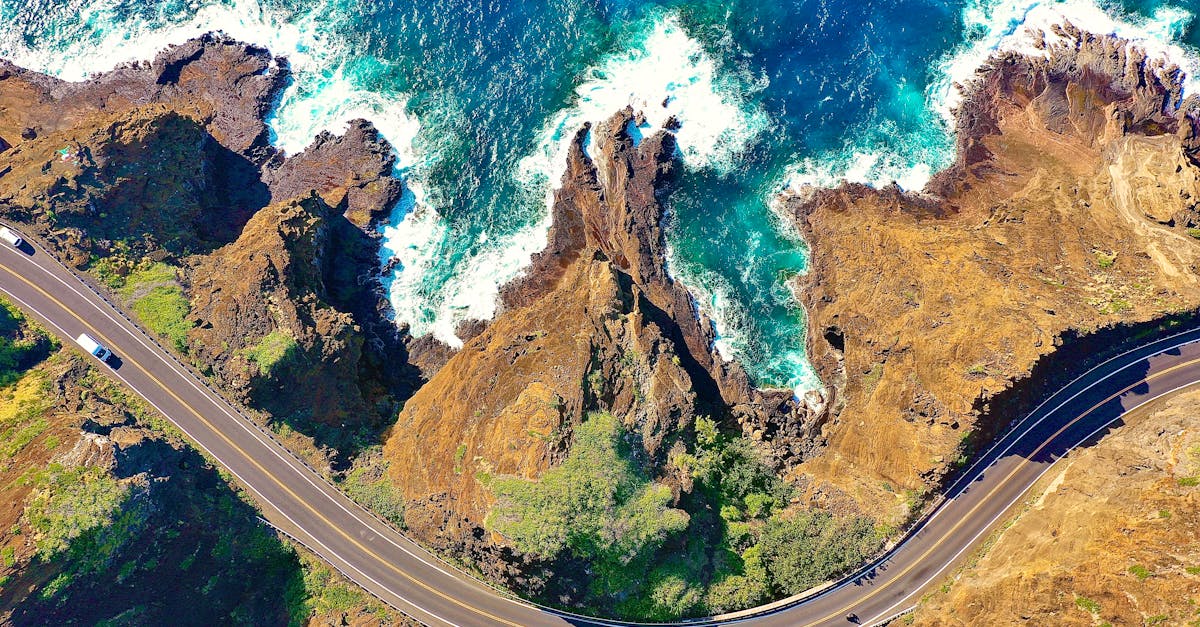 Aerial shot capturing a winding coastal road along a rugged, rocky shoreline with waves crashing into the cliffs.