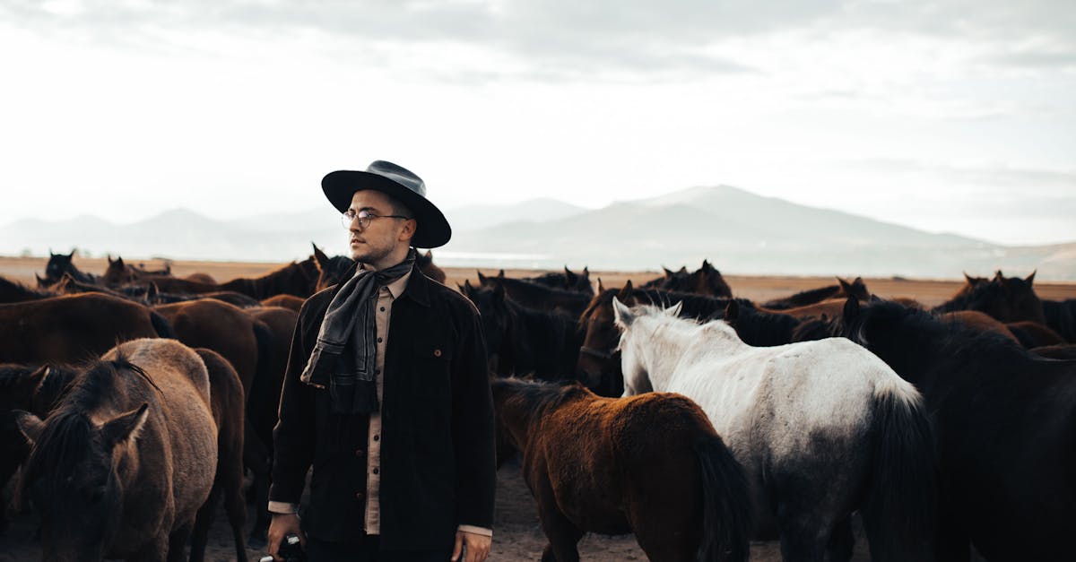 Man in cowboy hat among a herd of horses in a tranquil, open landscape.