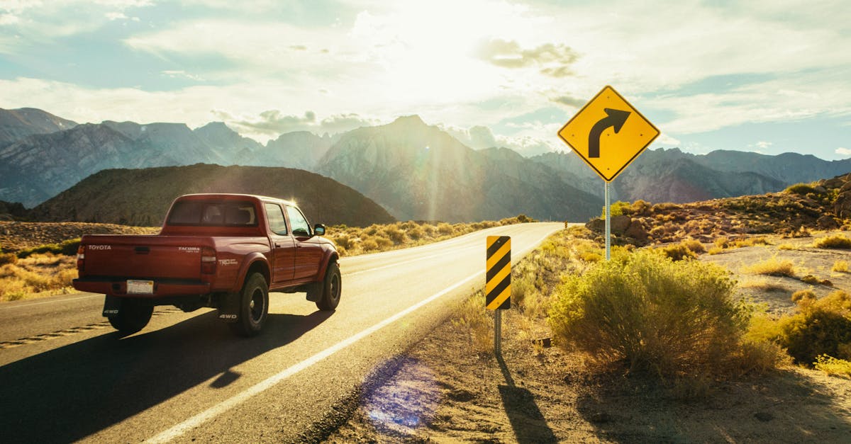 A red pickup truck driving on a sunlit road with mountains in the background and a road sign ahead.