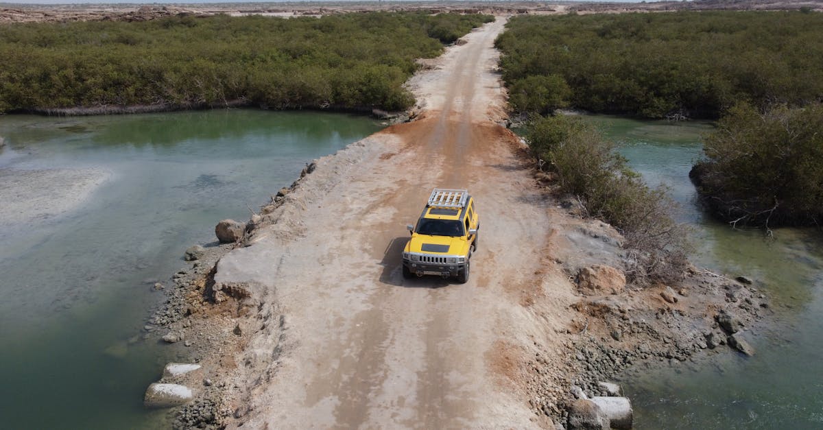 Aerial view of a yellow SUV crossing a remote desert road with surrounding greenery and water.
