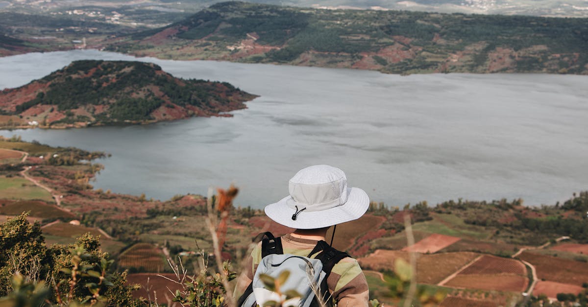 A hiker with a hat views a scenic lake and landscape from above, offering a tranquil outdoor experience.