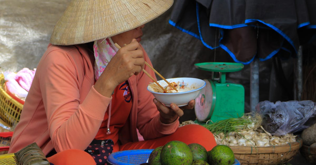A woman in a Vietnamese street market enjoying a meal while wearing a traditional hat.