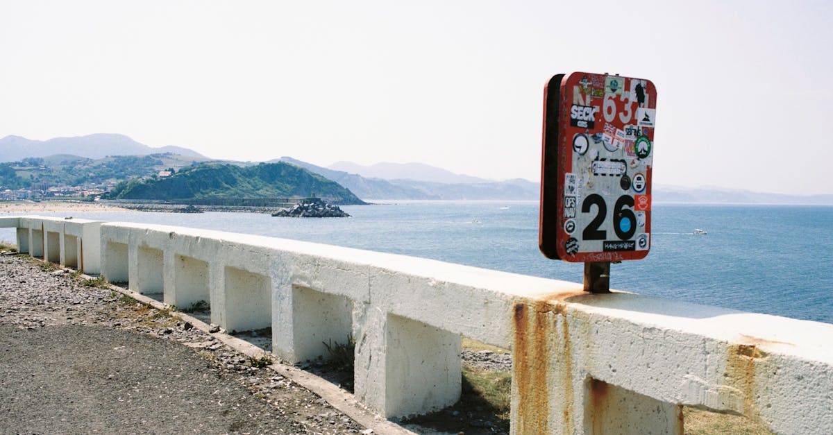 Graffiti-covered sign on a coastal road with ocean view and mountain backdrop.