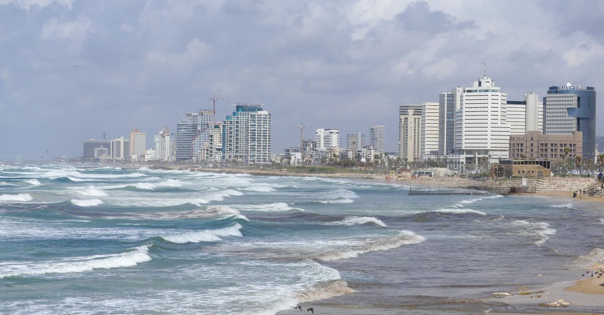 Waves crashing along the Tel Aviv shoreline with a view of modern skyscrapers under a cloudy sky.