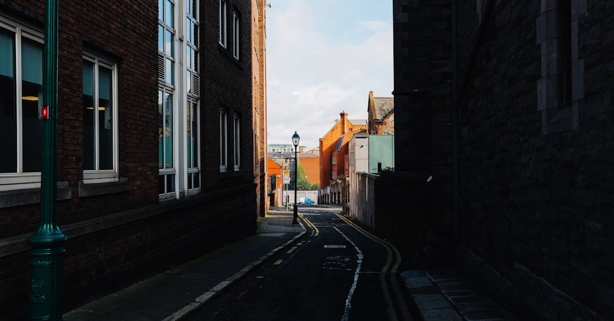 A narrow alleyway flanked by rustic brick buildings, leading to a sunlit street beyond.