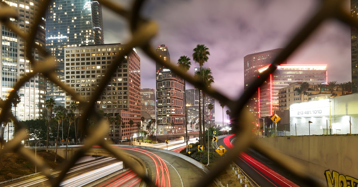 Long exposure night shot capturing the dynamic Los Angeles cityscape through a chain-link fence.