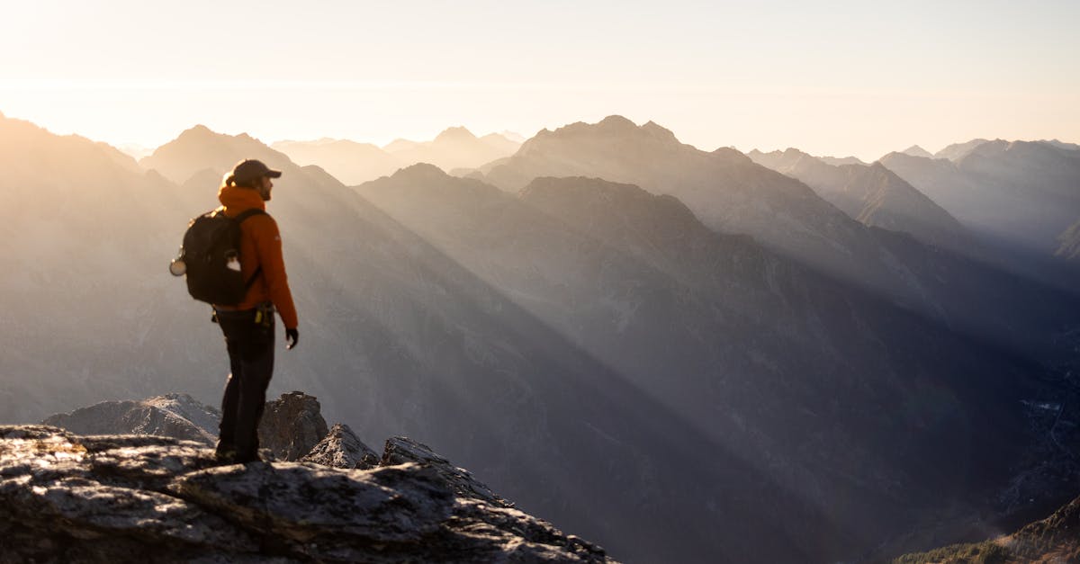 A solitary hiker in orange jacket stands on a rocky summit, admiring the sunrise over the mountain peaks.