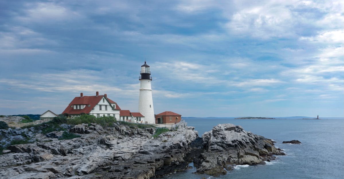 Scenic view of a historic lighthouse perched on rocky cliffs with a vast ocean backdrop and cloudy blue sky.