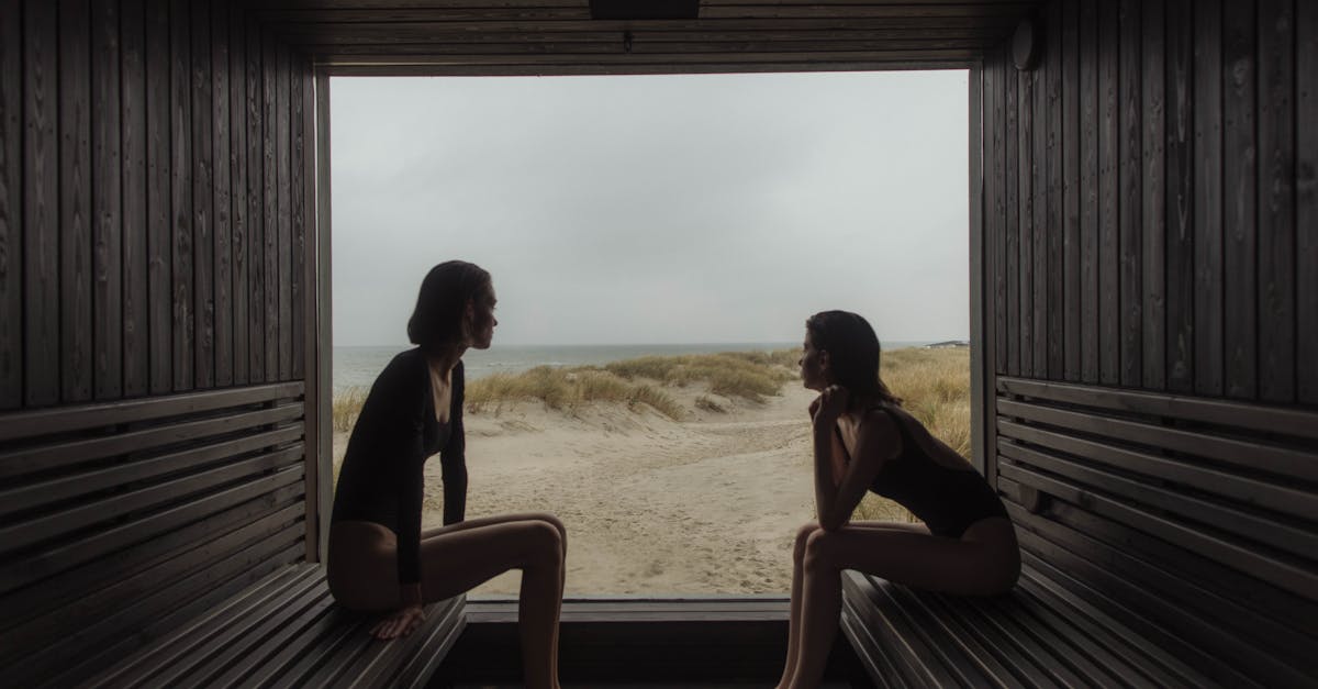 Two women in bathing suits sitting barefoot in a beach hut, looking out to the sandy coastline.