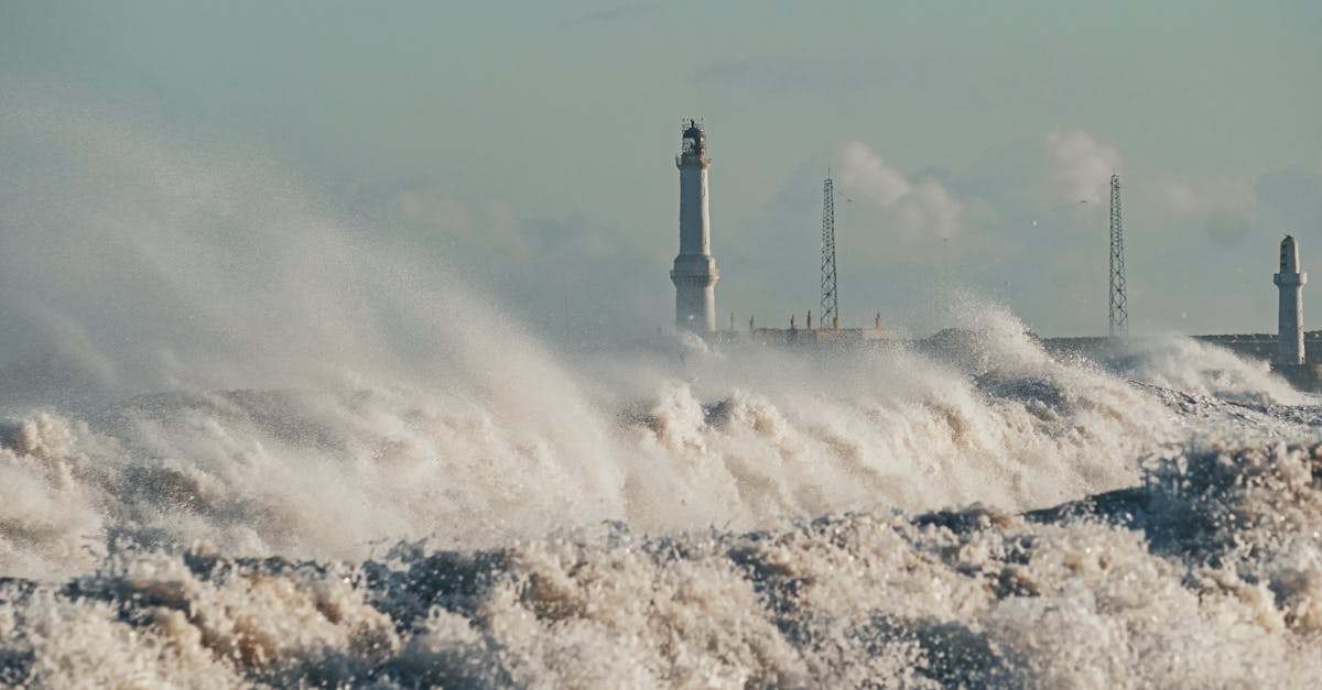 Powerful ocean waves crash near a lighthouse under a clear sky, showcasing nature's force.
