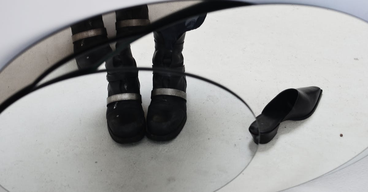 Crop anonymous person feet in black boots with decorative metal details standing near black stylish shoe and reflecting in oval mirror