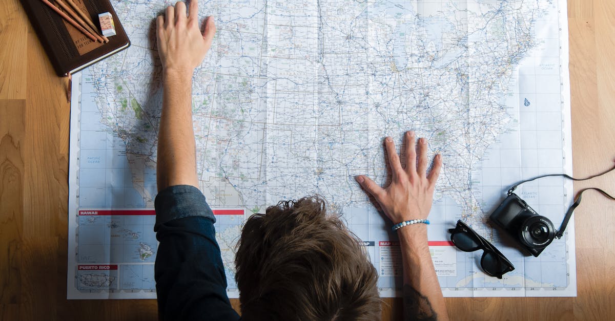 Top view of anonymous young male in casual clothes sitting on wooden floor with photo camera and coffee cup and pointing on paper map while planning trip route