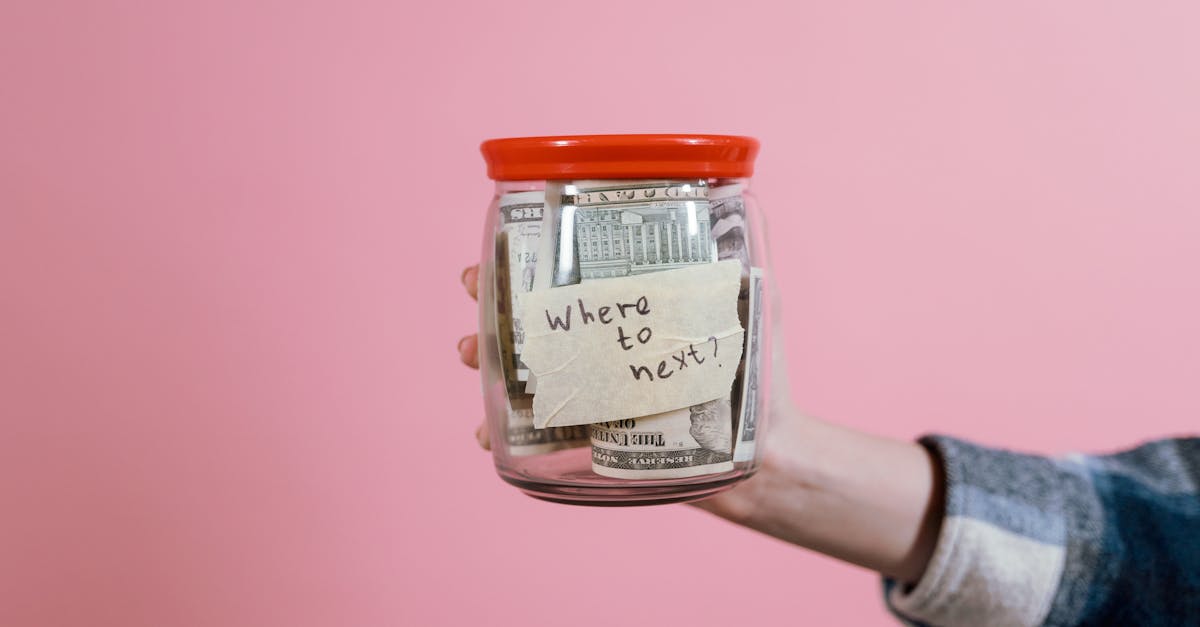 A hand holding a glass jar filled with dollar bills labeled 'Where to next?' against a pink background, symbolizing savings and future planning.