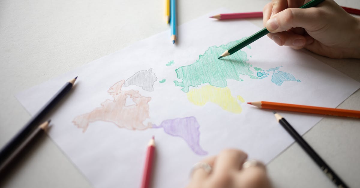 Crop anonymous person with colored pencils coloring continents on white paper on table with various scattered stationery in light room
