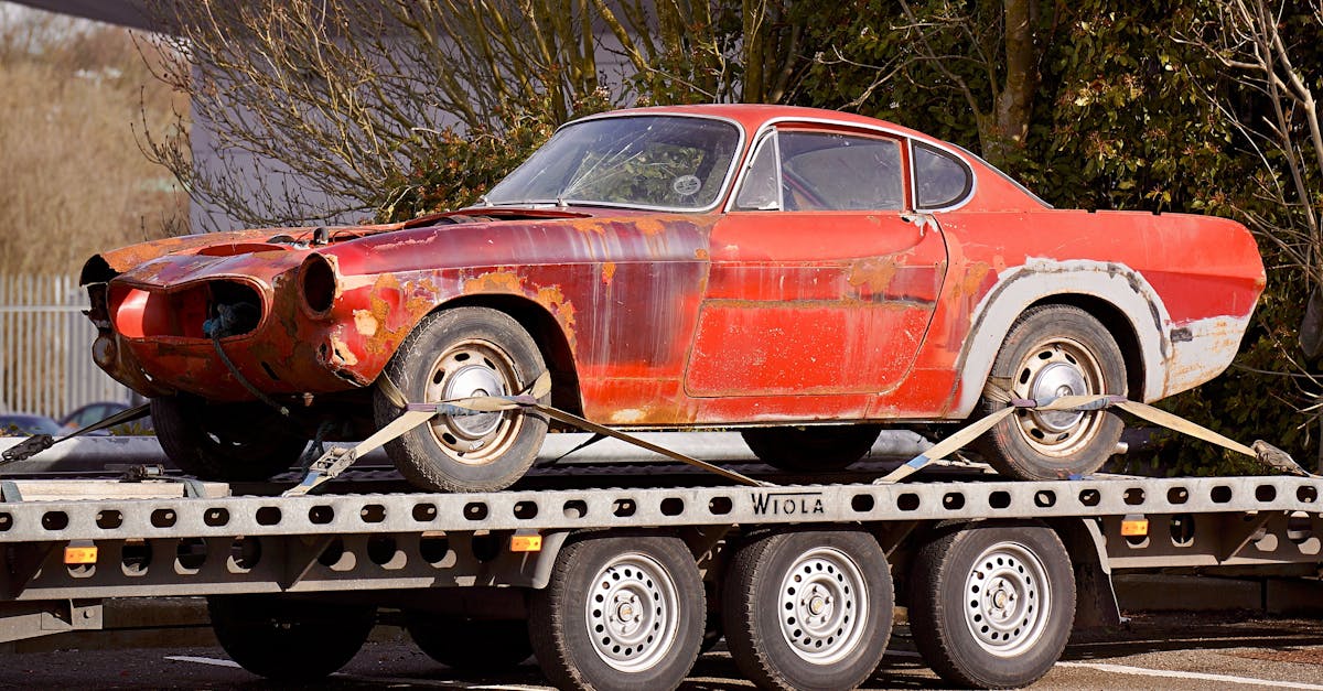 A worn-out, rusty vintage car secured on a flatbed trailer, outdoors under daylight.